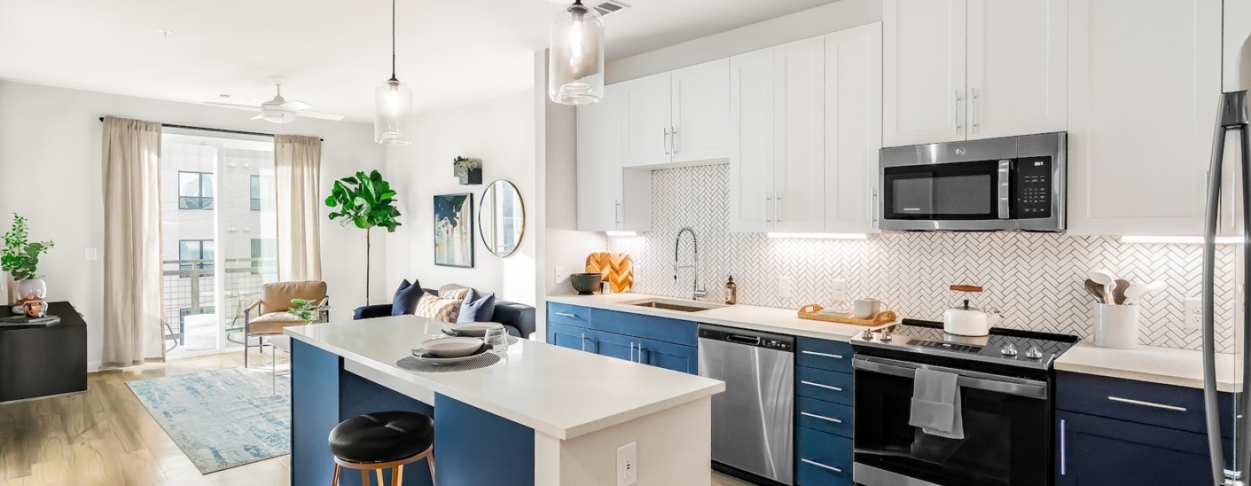 Modern apartment kitchen with white upper cabinets, blue lower cabinets, stainless steel appliances, island seating, and open living area with large windows.