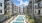 Courtyard view of a modern apartment complex featuring multiple balconies and a central pool area with in-water lounge chairs, surrounded by sun loungers and greenery