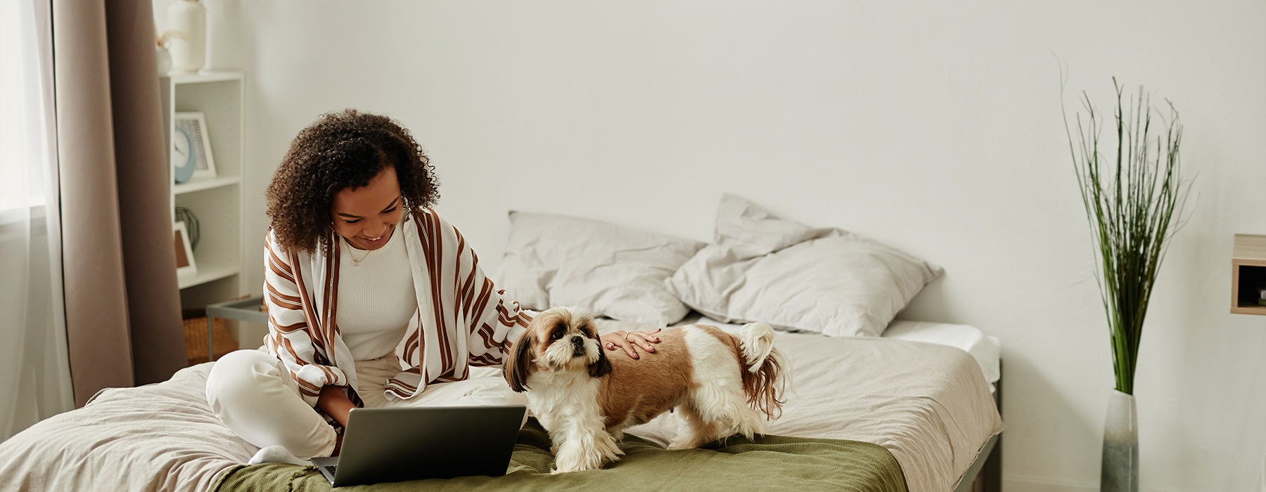 A woman multitasking, petting her dog while working on her laptop while seated on her bed.
