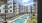 View of a modern apartment complex courtyard featuring a large central pool with lounge chairs, surrounded by balconies, greenery, and poolside seating areas