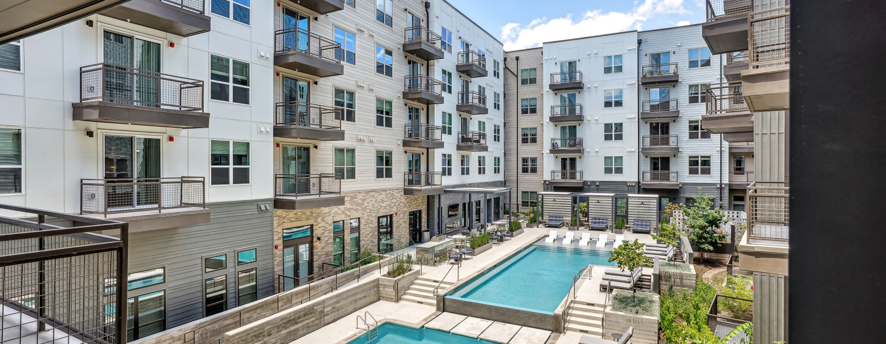 A modern apartment complex courtyard features two pools, lounge chairs, greenery, and surrounding buildings with balconies under a bright sky