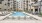 Modern outdoor pool area with in-water lounge chairs, surrounded by sun loungers and a pergola, with a multi-story apartment building in the background