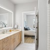 Modern bathroom with a large illuminated mirror, wooden vanity with dual sinks, glass-enclosed shower, and a doorway leading to a bedroom with a neatly made bed