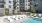 Poolside area featuring in-water lounge chairs, a pergola with cushioned seating, and additional sun loungers, surrounded by modern apartment buildings and planters with greenery