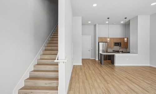 Modern kitchen with wood flooring, stainless steel appliances, pendant lights, and a staircase leading upstairs